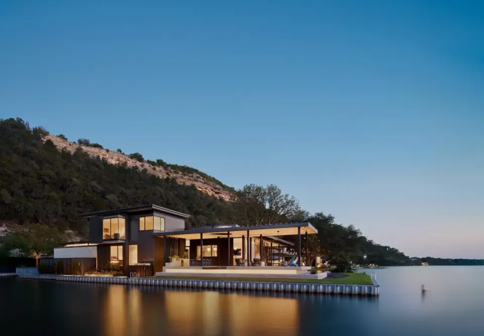 Lakeside modern home at dusk, glowing pavilion over the water with a hill behind